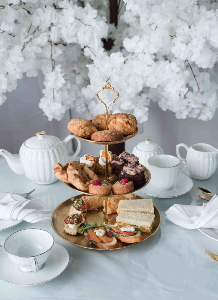 A three-tiered stand of afternoon tea treats on a table with a white teapot and floral arrangement in the background.