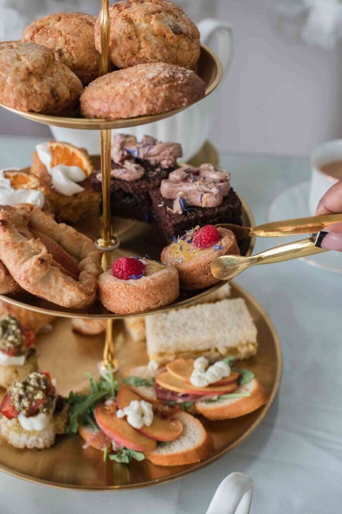 A close-up shot of a three-tiered platter with a variety of afternoon tea treats, including scones, sandwiches, and desserts. A hand with gold tongs is reaching for a pastry.