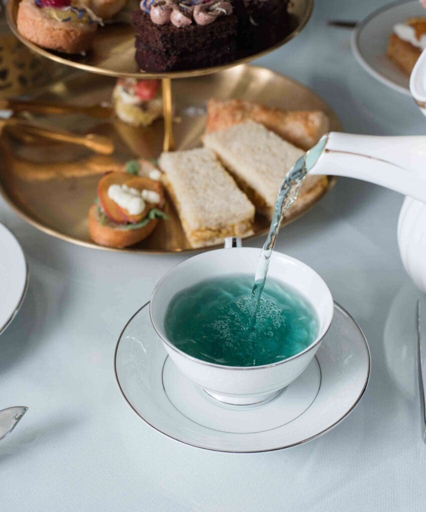 A hand pouring vibrant blue tea from a white teapot into a teacup, with a tiered platter of treats in the background.