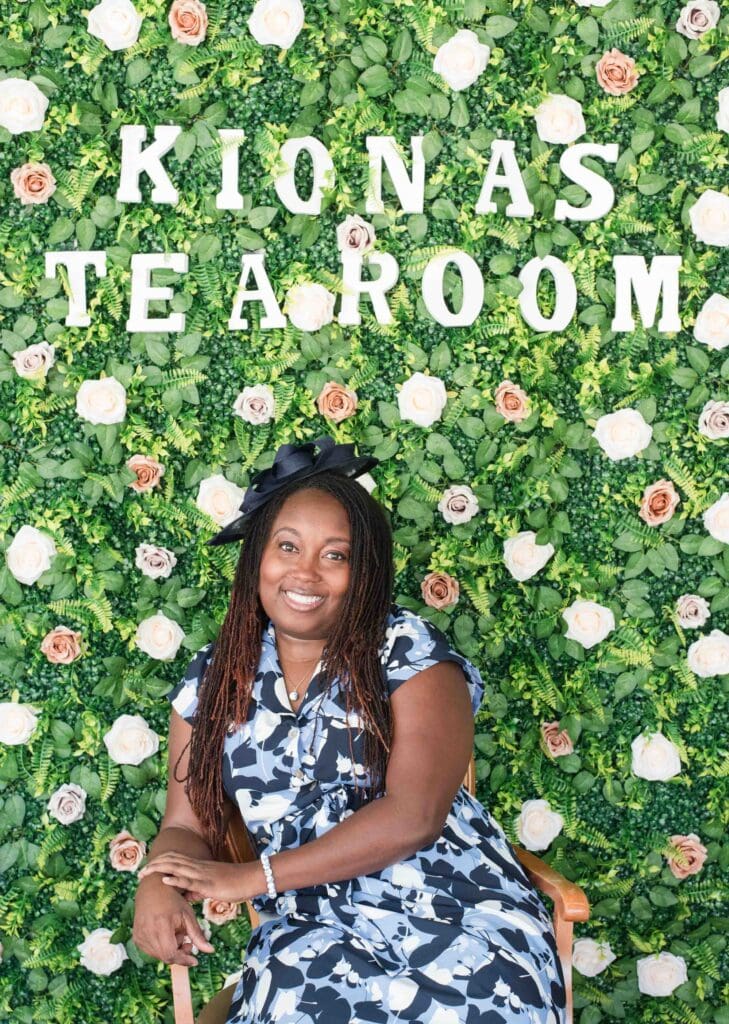 Tea room marketing photos for Kiona's Tea Room. A smiling woman sits in a chair in front of a green wall covered in white and pink flowers, with the words "KIONAS TEA ROOM" spelled out in white letters above her.
